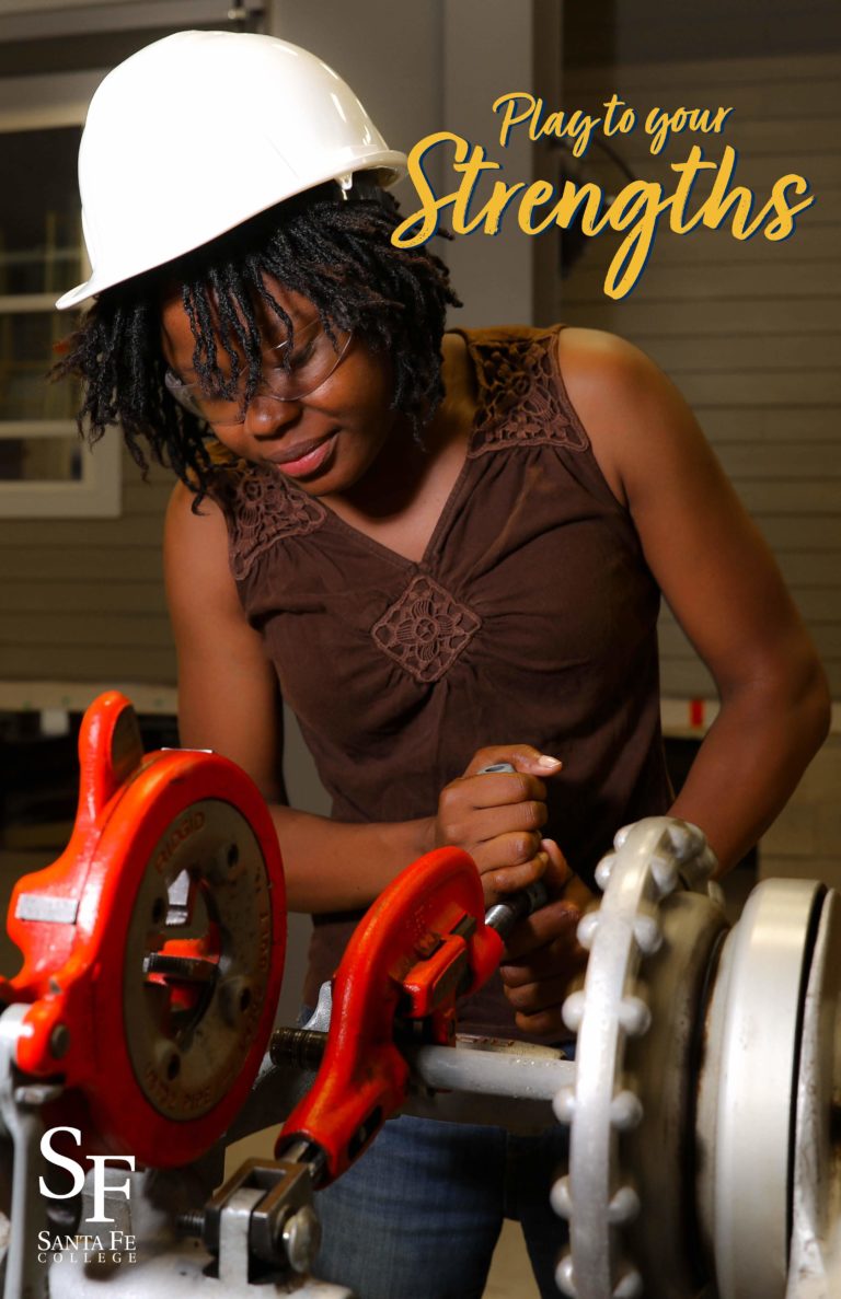 A young woman with dark skin and black braids is wearing blue jeans, a tank top and a white hardhat. She's operating a large, bright orange pipe cutting tool.